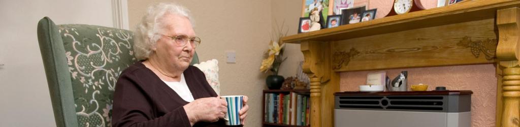 Older lady in armchair drinking tea in front of gas fire